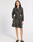 A woman modeling a black long-sleeve mini dress with a colorful floral and botanical print, featuring a tie waist belt and collar, paired with dark olive green loafers against a light gray studio background.