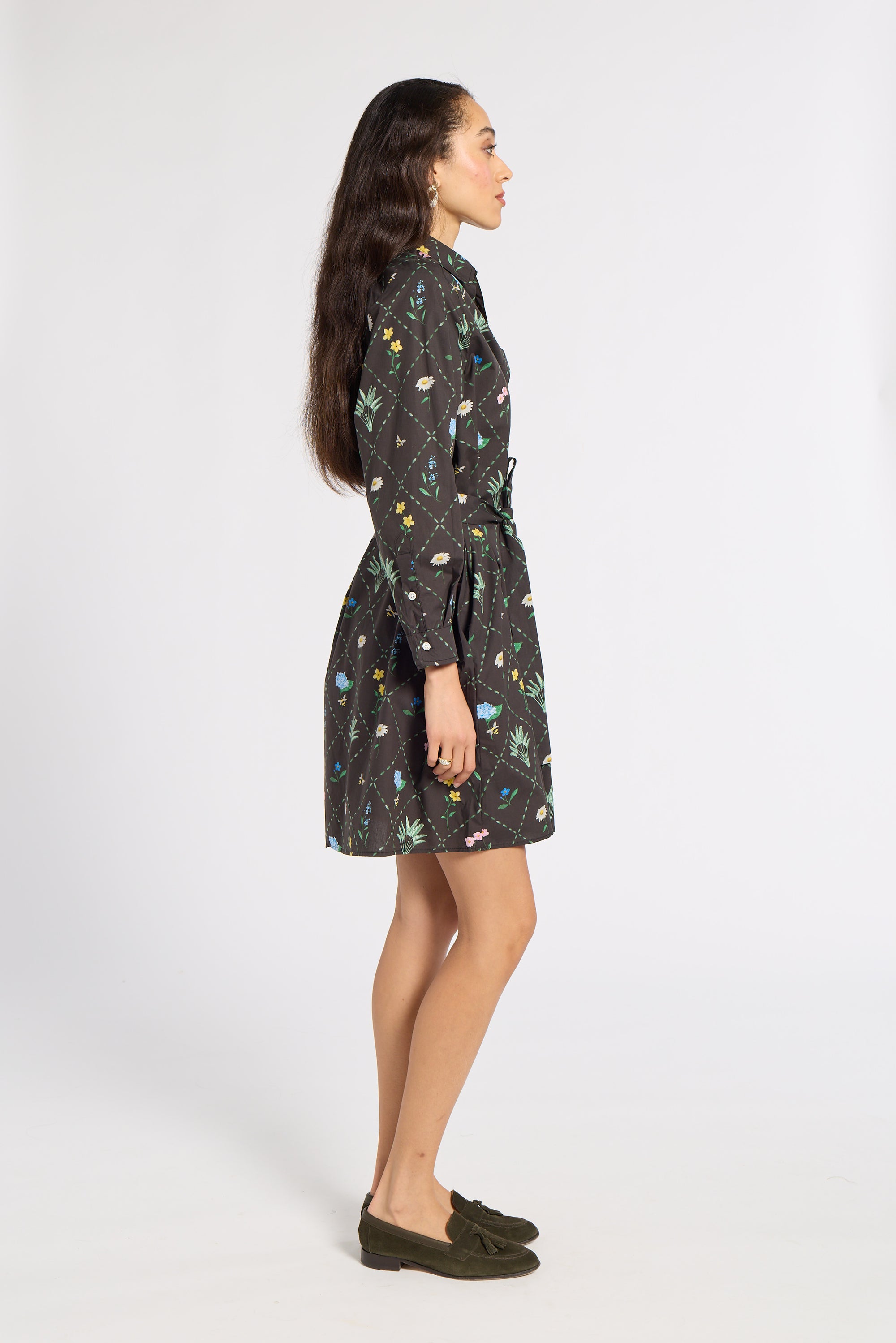 A woman shown in profile wearing a dark black wrap dress with a colorful, whimsical all-over print. She is wearing olive green loafers and standing against a plain white studio background.