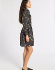 A woman shown in profile wearing a dark black wrap dress with a colorful, whimsical all-over print. She is wearing olive green loafers and standing against a plain white studio background.