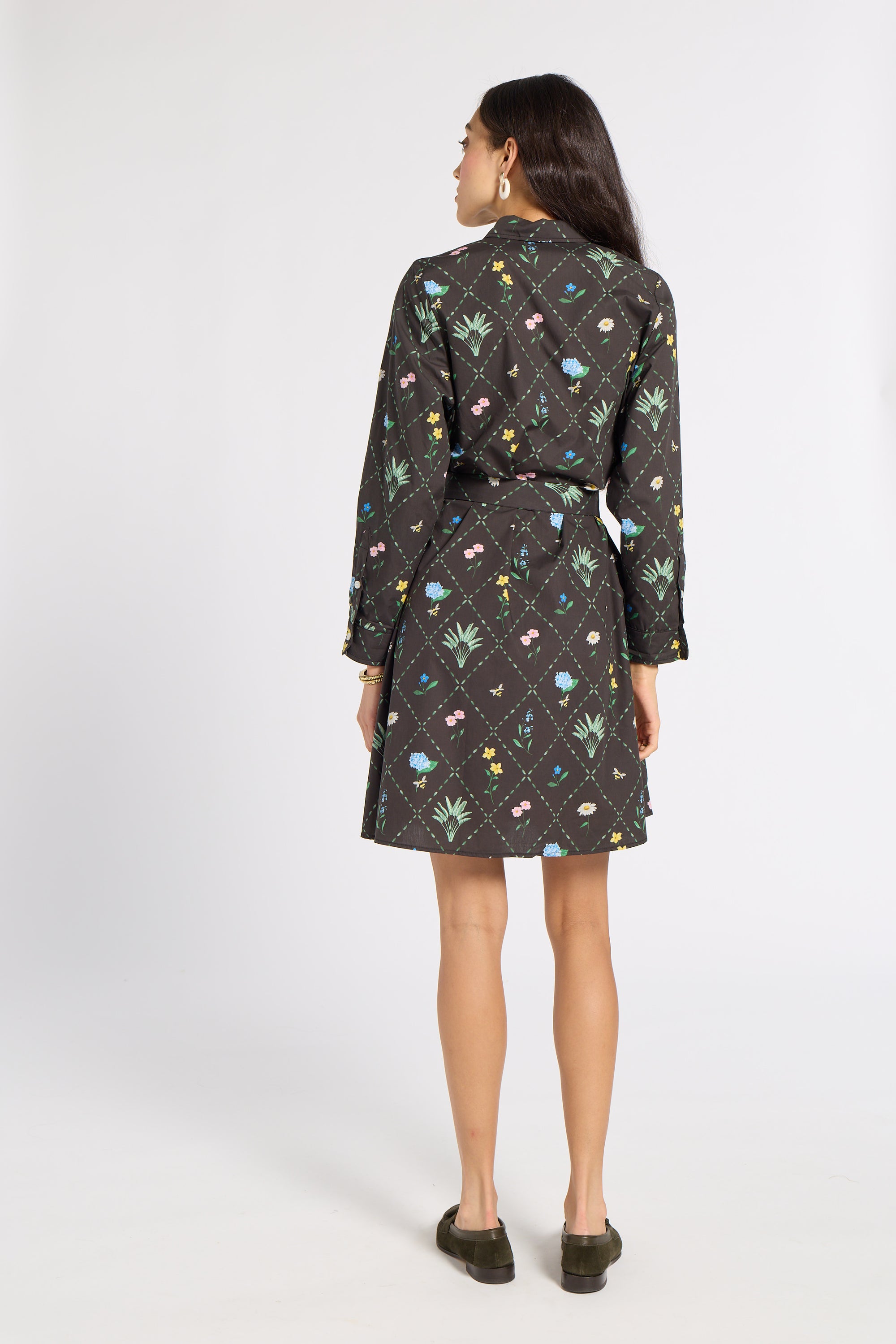 A woman viewed from the back wearing a black belted shirt dress with a colorful floral and butterfly pattern, paired with dark olive slip-on shoes, against a light gray studio background.