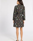A woman viewed from the back wearing a black belted shirt dress with a colorful floral and butterfly pattern, paired with dark olive slip-on shoes, against a light gray studio background.