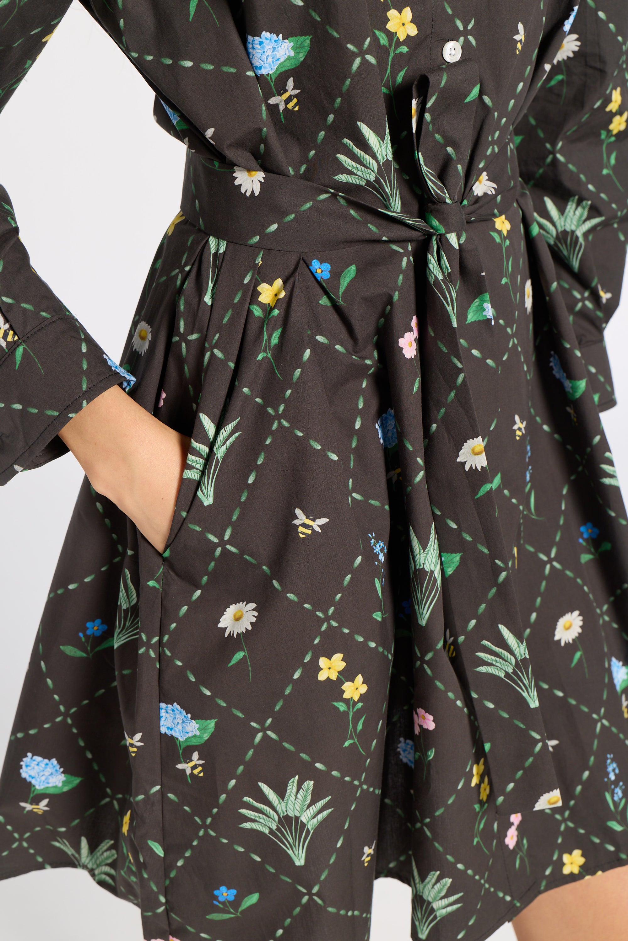 A close-up of a black dress with a green grid pattern and colorful embroidered wildflowers, featuring a tie waist and a hand tucked into a pocket.
