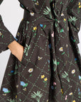 A close-up of a black dress with a green grid pattern and colorful embroidered wildflowers, featuring a tie waist and a hand tucked into a pocket.