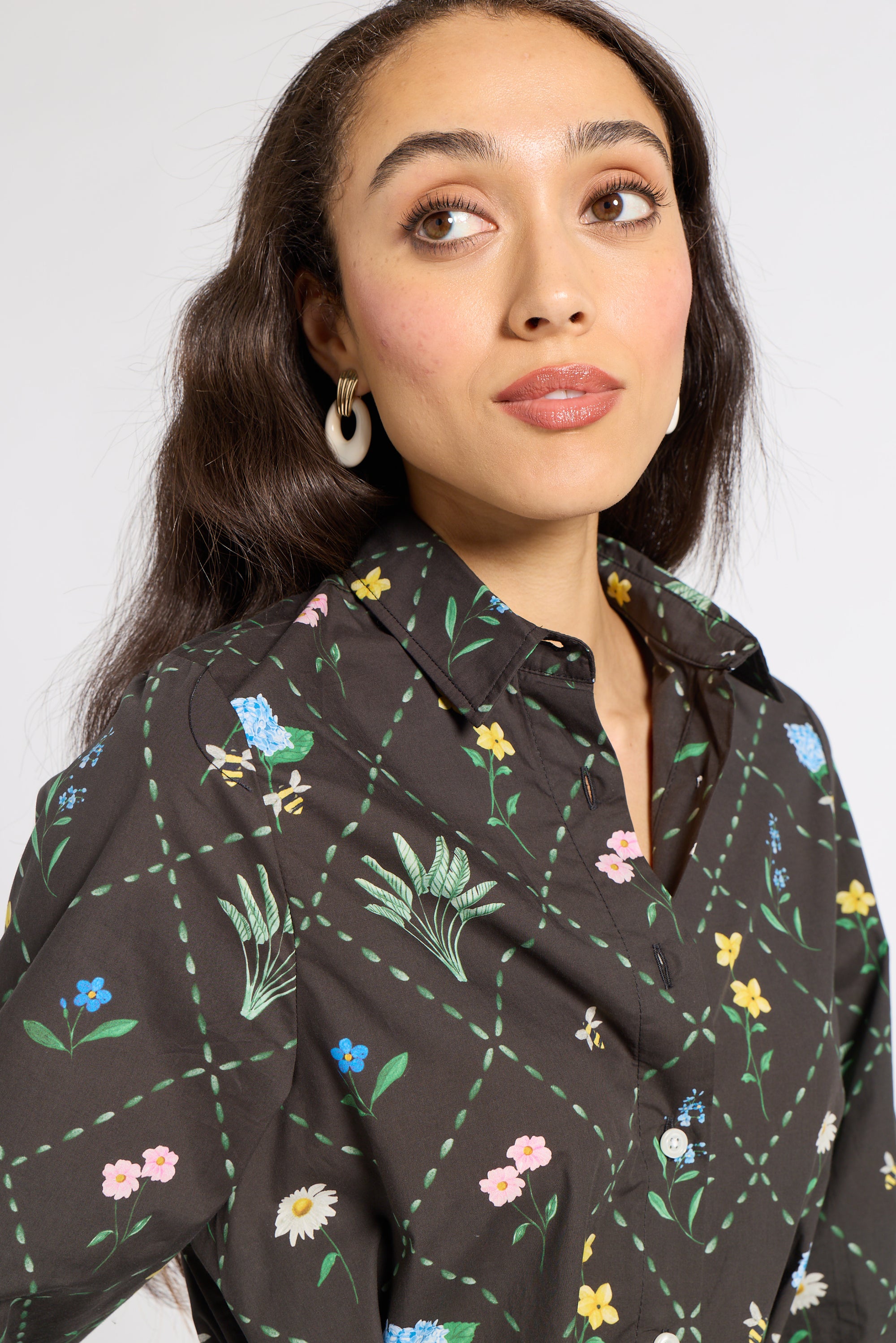 A woman wearing a black button-up shirt with a colorful floral and bee pattern, paired with white hoop earrings, photographed against a light gray background.