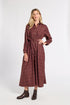 A woman modeling a long-sleeved, button-front midi dress in a dark reddish-brown color with a subtle windowpane plaid pattern. The dress features a matching self-tie belt at the waist and is styled with dark brown leather ankle boots against a plain white studio background.