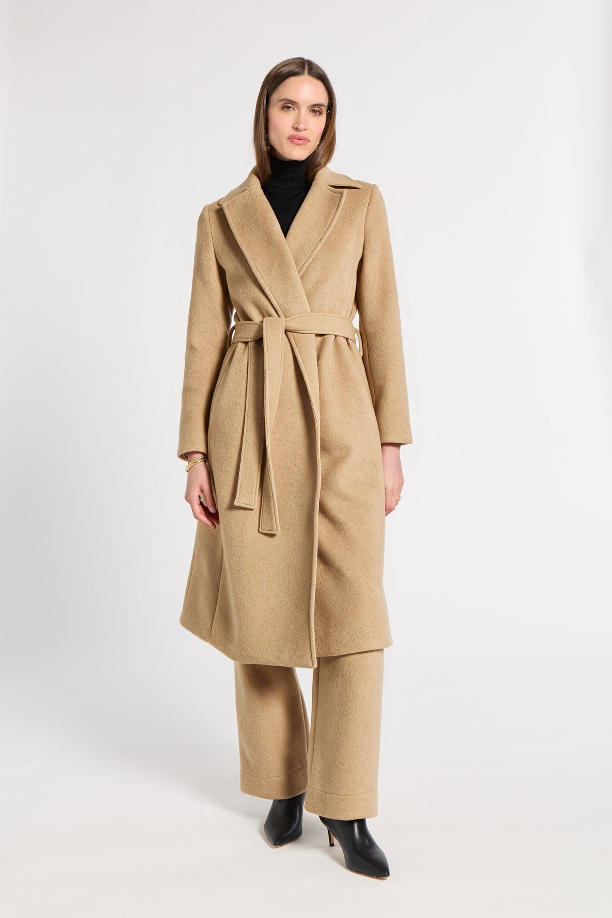 A woman modeling a long camel-colored cashmere coat with a matching belt and wide-leg trousers, styled with a black turtleneck and black pointed-toe heels against a plain white studio background.