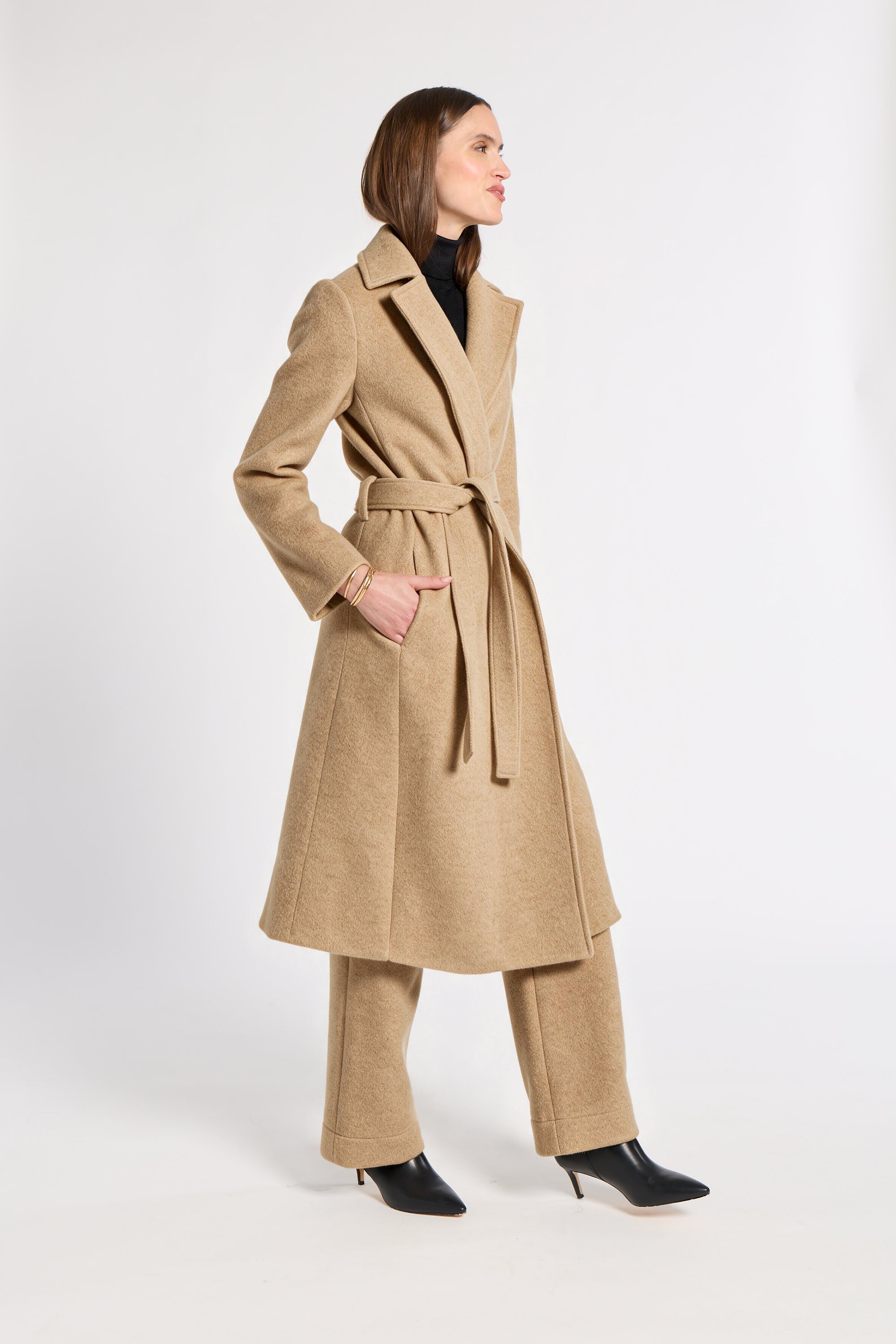 A woman modeling a long camel-colored belted coat with matching wide-leg trousers and black pointed-toe heels, photographed against a plain light gray studio background.