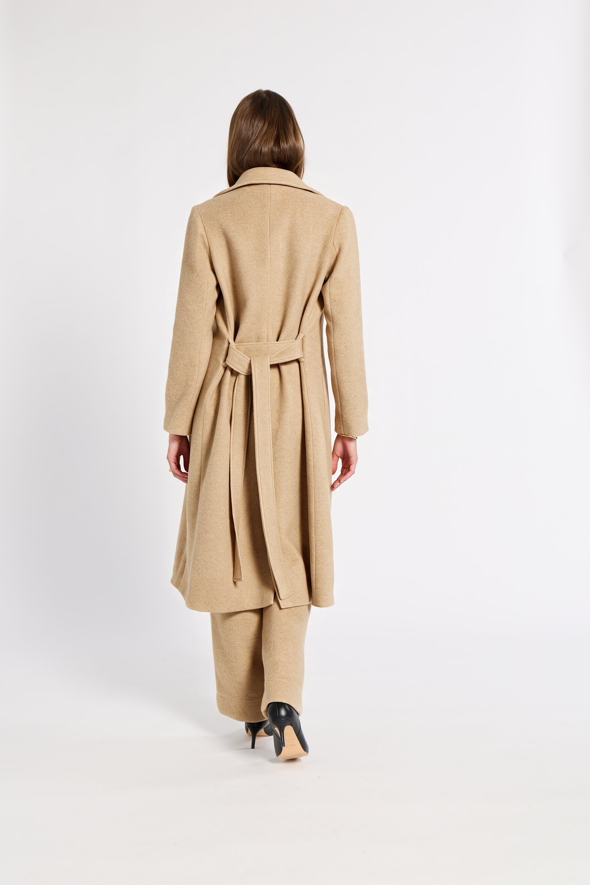 A woman shown from the back wearing a long camel-colored belted coat and matching trousers, standing against a plain white studio background.
