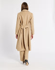 A woman shown from the back wearing a long camel-colored belted coat and matching trousers, standing against a plain white studio background.