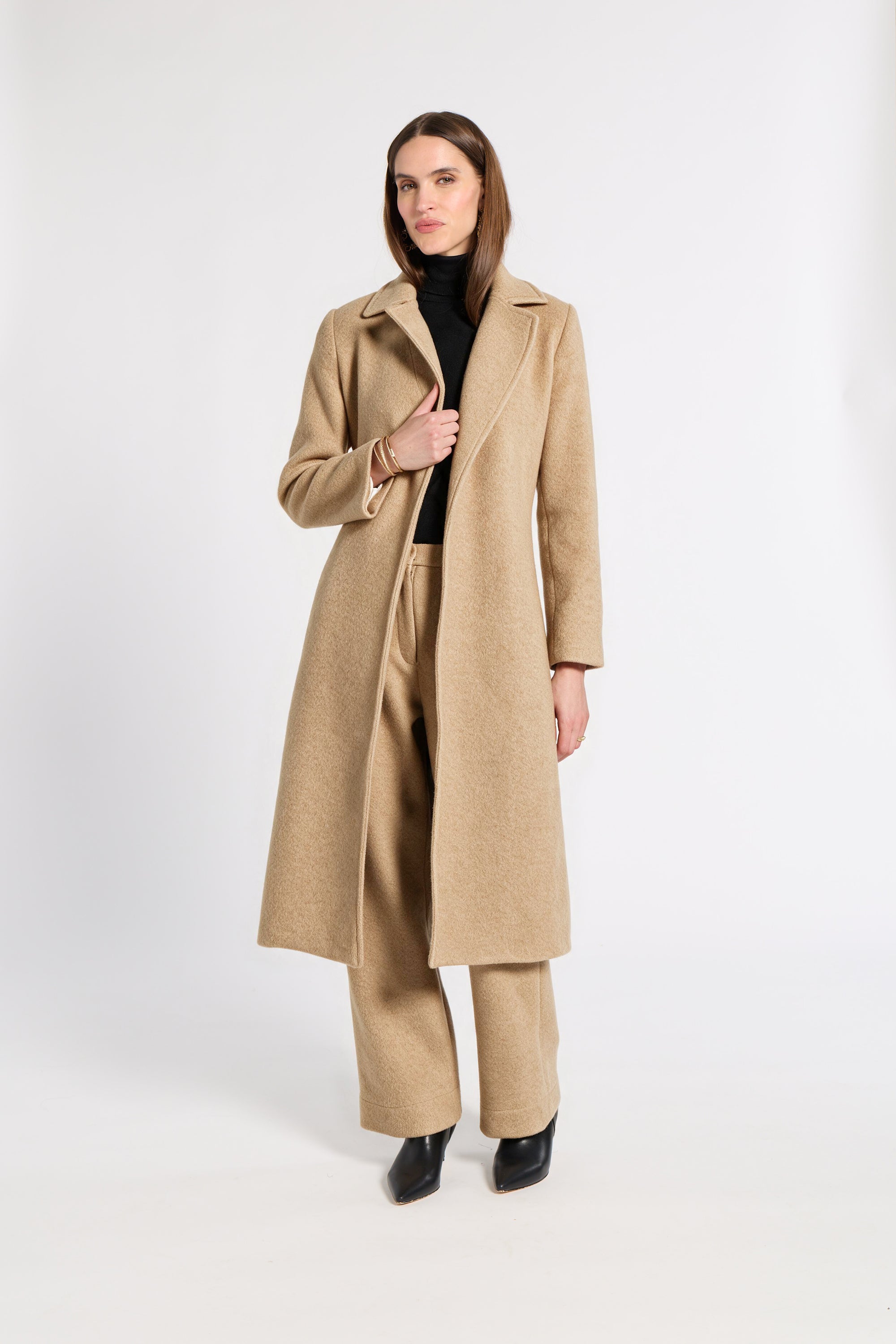 A woman modeling a long camel-colored cashmere coat over a black turtleneck and matching wide-leg trousers, standing against a plain white studio background.