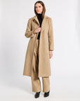 A woman modeling a long camel-colored cashmere coat over a black turtleneck and matching wide-leg trousers, standing against a plain white studio background.