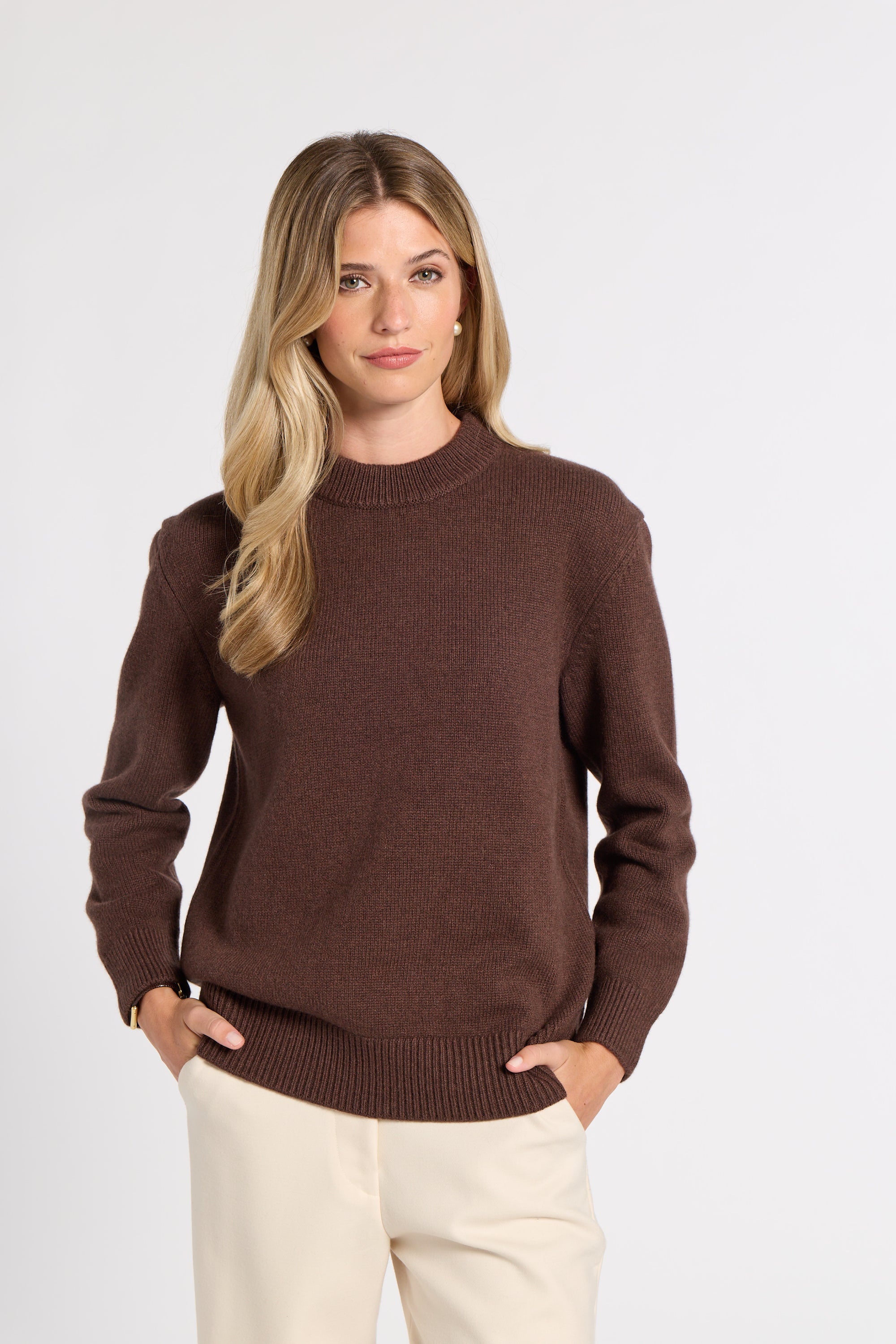 A woman modeling a dark brown crewneck knit sweater with cream pants, standing against a light gray studio background.