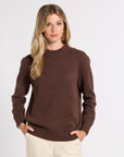 A woman modeling a dark brown crewneck knit sweater with cream pants, standing against a light gray studio background.