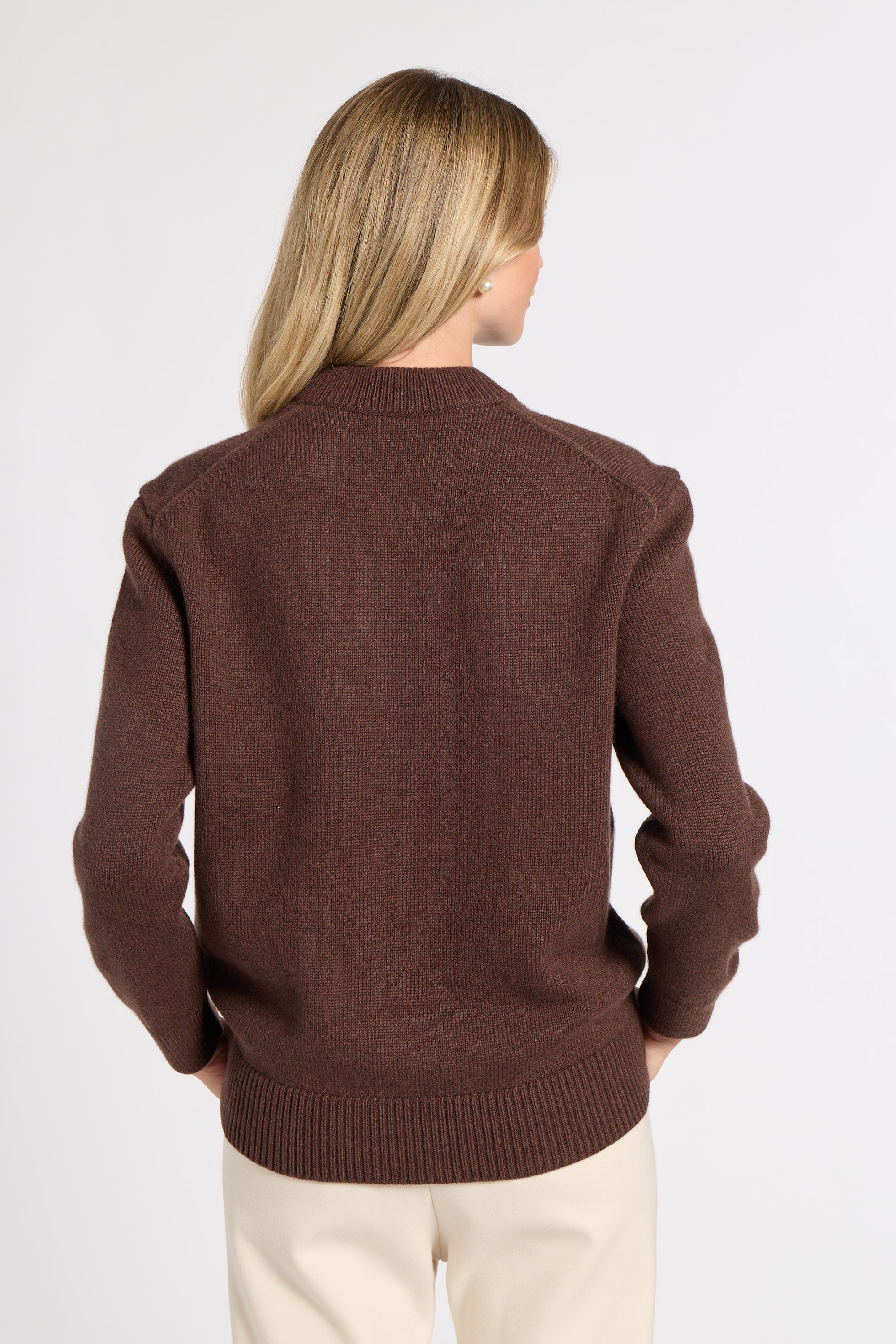 A woman viewed from the back wearing a brown knit sweater and cream pants against a plain light gray studio background.