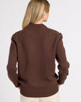 A woman viewed from the back wearing a brown knit sweater and cream pants against a plain light gray studio background.