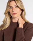 A blonde woman wearing a brown ribbed knit crewneck sweater and small pearl stud earrings, posing against a light gray studio background.