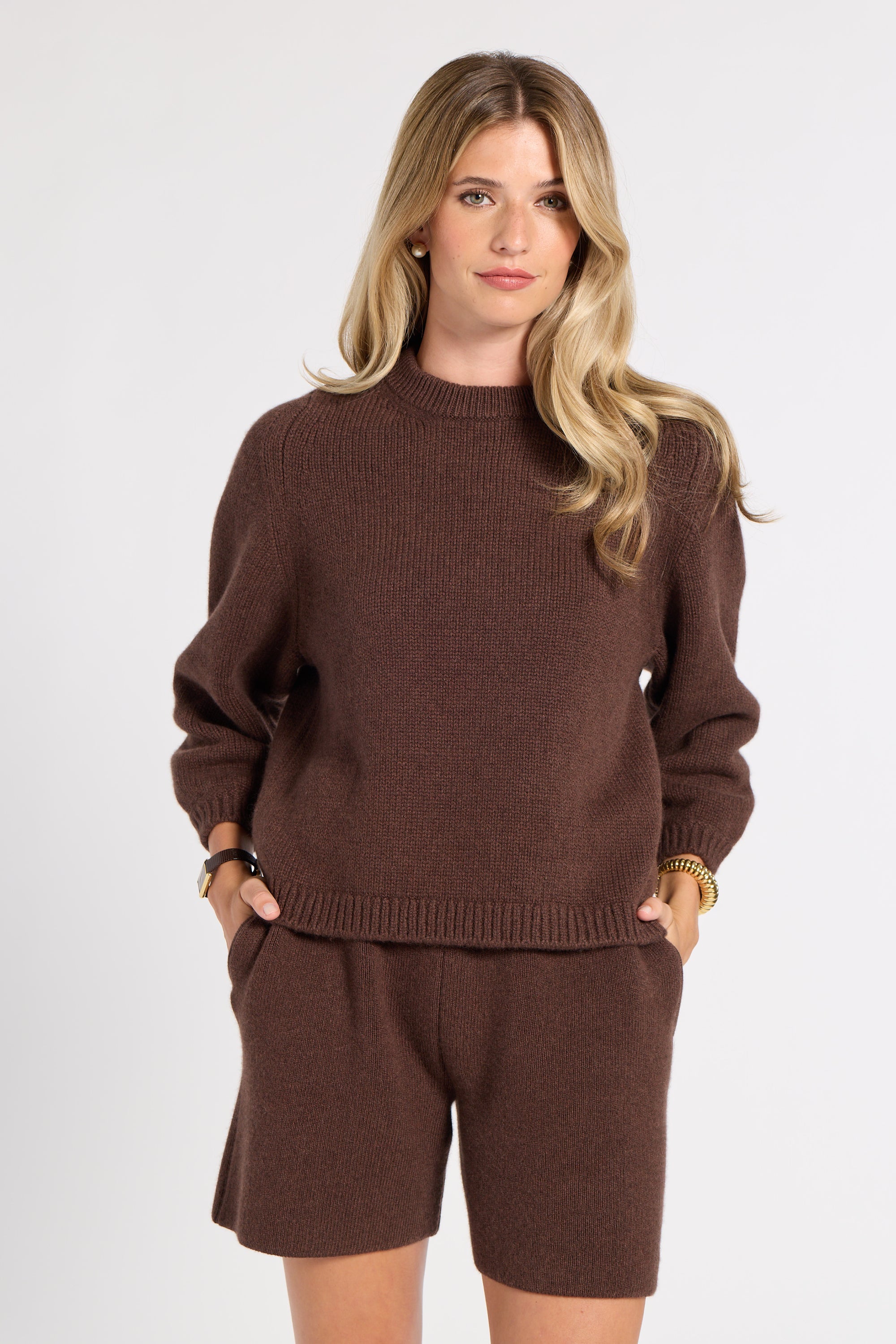 A woman modeling a matching brown knit sweater and shorts set against a plain light gray studio background.