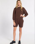 A woman modeling a matching brown knit sweater and shorts set, paired with black leather shoes, against a plain white studio background.