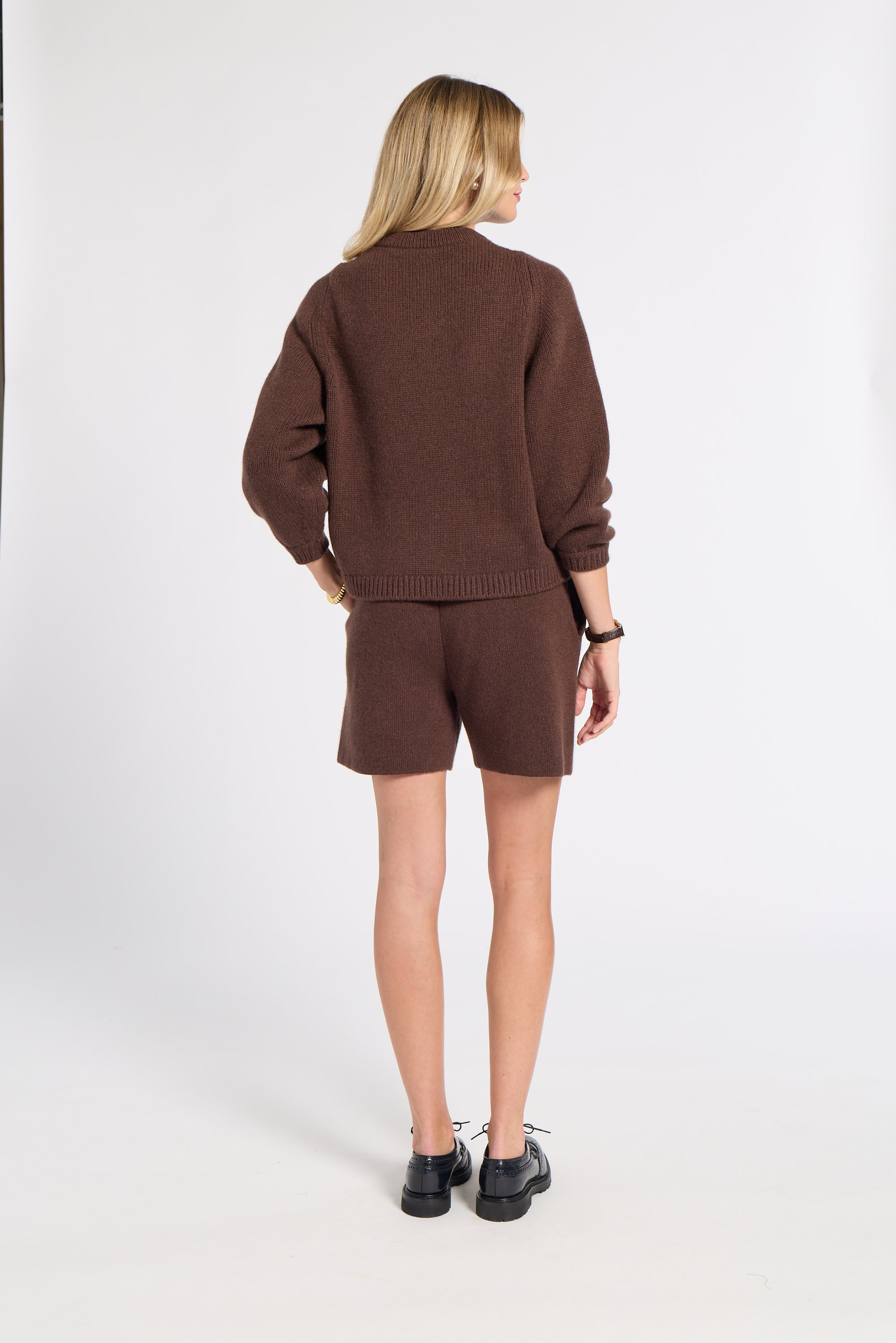 A woman is shown from behind wearing a matching brown knit sweater and shorts set, paired with black shoes, against a plain white studio background.