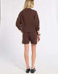 A woman is shown from behind wearing a matching brown knit sweater and shorts set, paired with black shoes, against a plain white studio background.