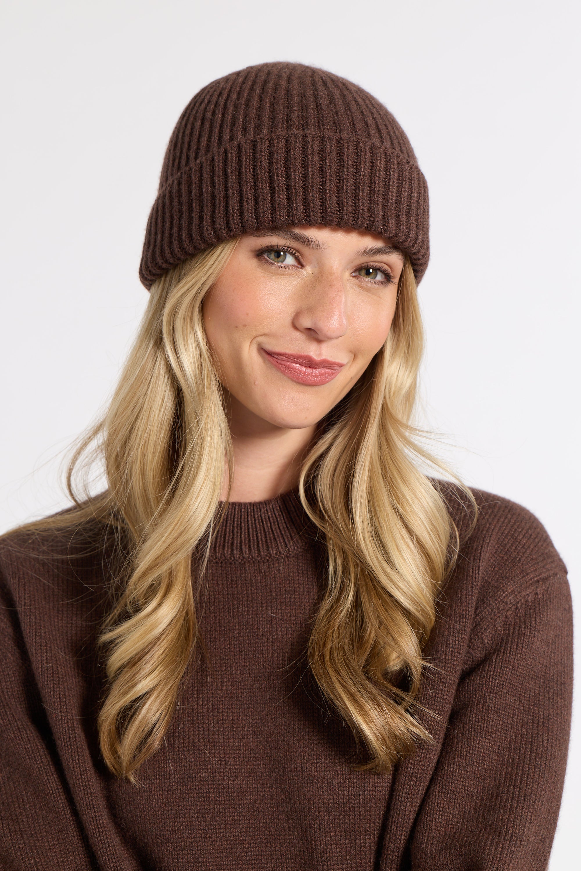 A woman with long blonde hair wearing a ribbed brown beanie and a matching brown knit sweater, photographed against a clean light gray studio background.