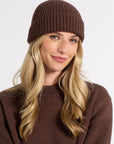 A woman with long blonde hair wearing a ribbed brown beanie and a matching brown knit sweater, photographed against a clean light gray studio background.