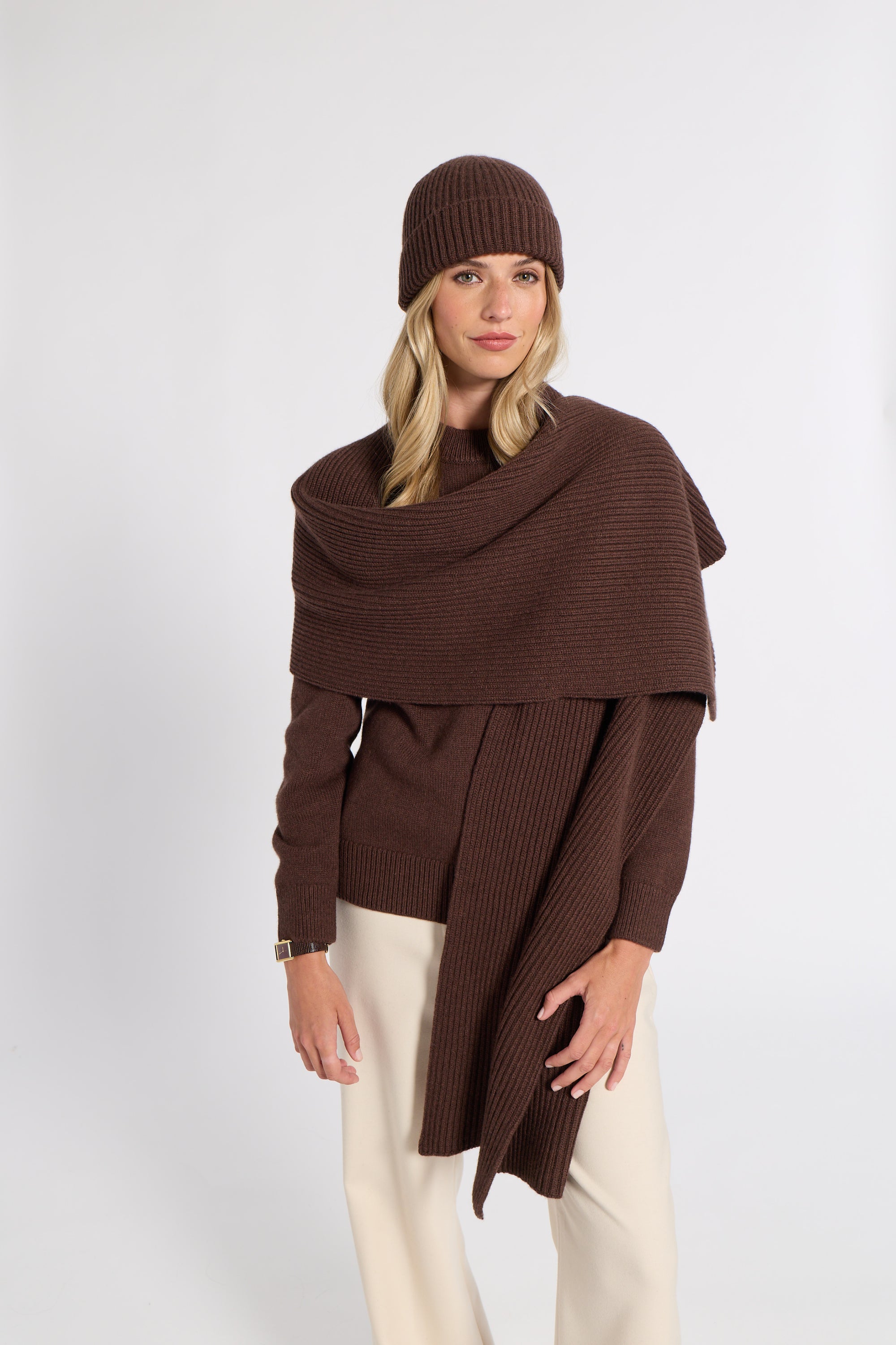 A woman modeling a dark brown ribbed knit wrap sweater, matching beanie, and cream pants against a light gray studio background.