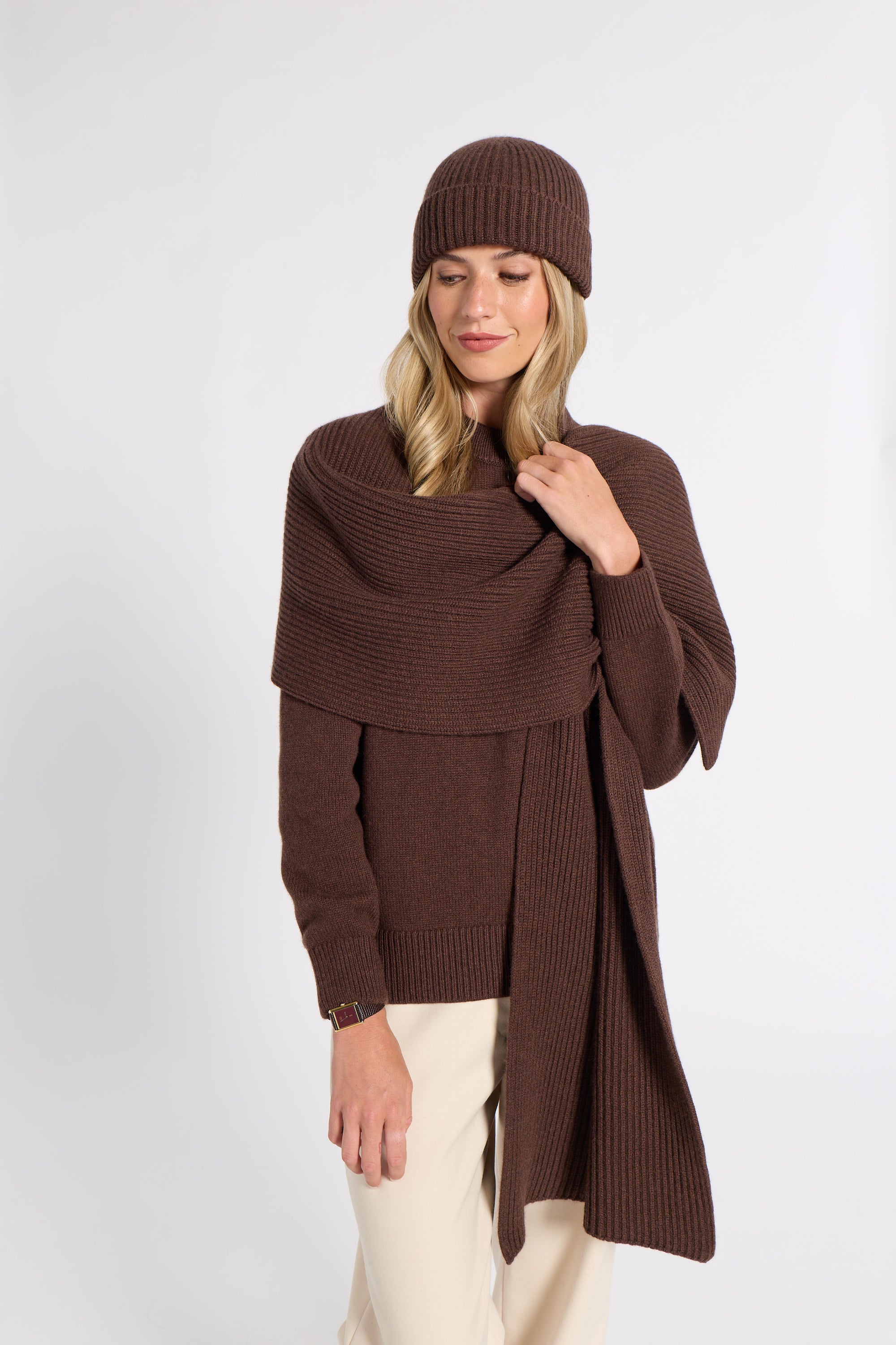A woman modeling a brown ribbed knit beanie, matching wrap shawl, and long-sleeve poncho-style sweater, paired with cream pants, against a plain light gray studio background.