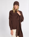 A woman modeling a brown ribbed knit beanie, matching wrap shawl, and long-sleeve poncho-style sweater, paired with cream pants, against a plain light gray studio background.