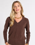 A blonde woman wearing a brown V-neck knit sweater, cream pants, and gold jewelry, standing against a light gray studio background.
