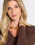 A blonde woman wearing a chunky gold chain necklace and a matching gold bangle bracelet, paired with a brown V-neck knit sweater against a light gray background.