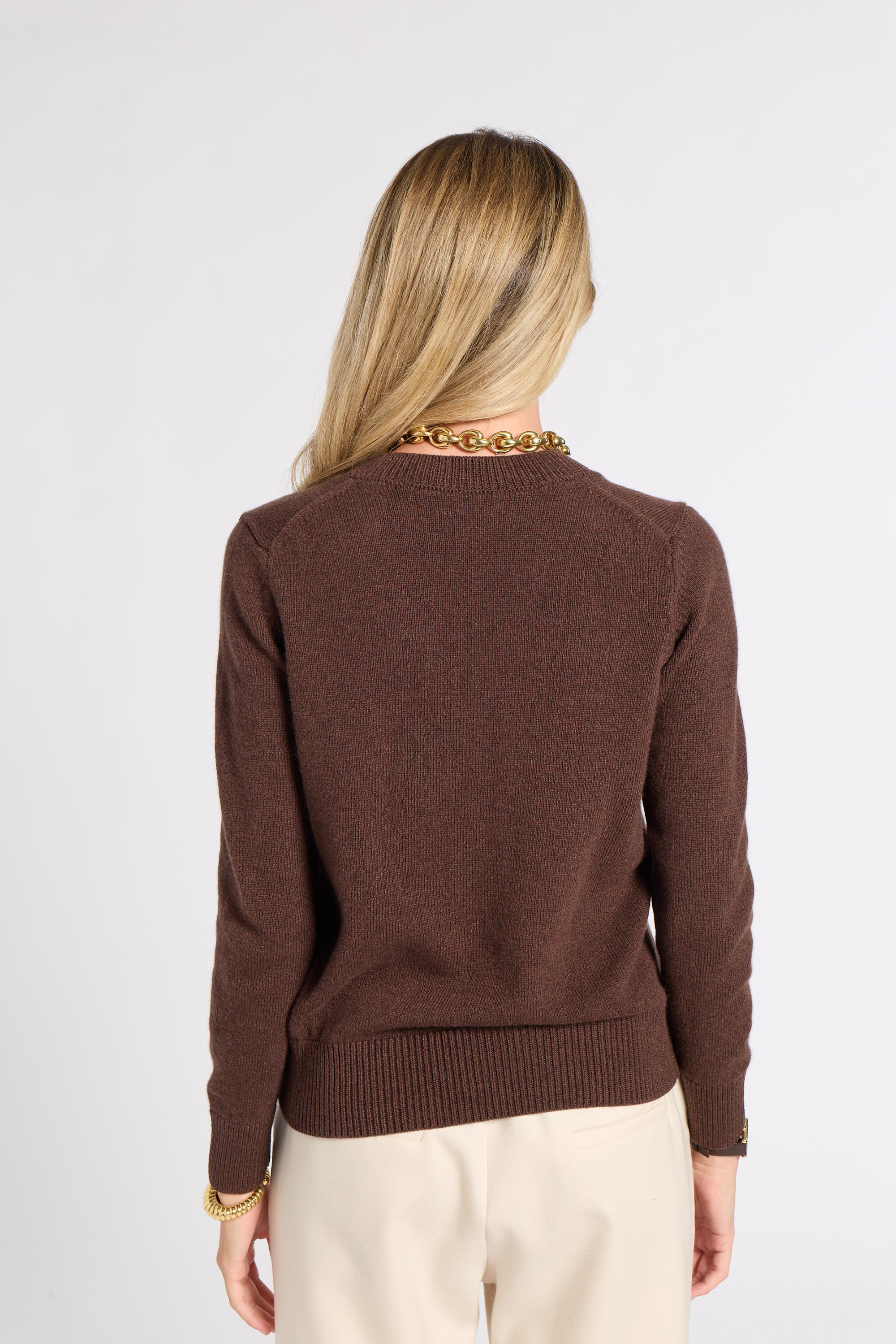 A woman viewed from the back wearing a brown knit sweater and cream trousers, standing against a plain light gray studio background.