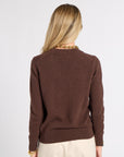 A woman viewed from the back wearing a brown knit sweater and cream trousers, standing against a plain light gray studio background.