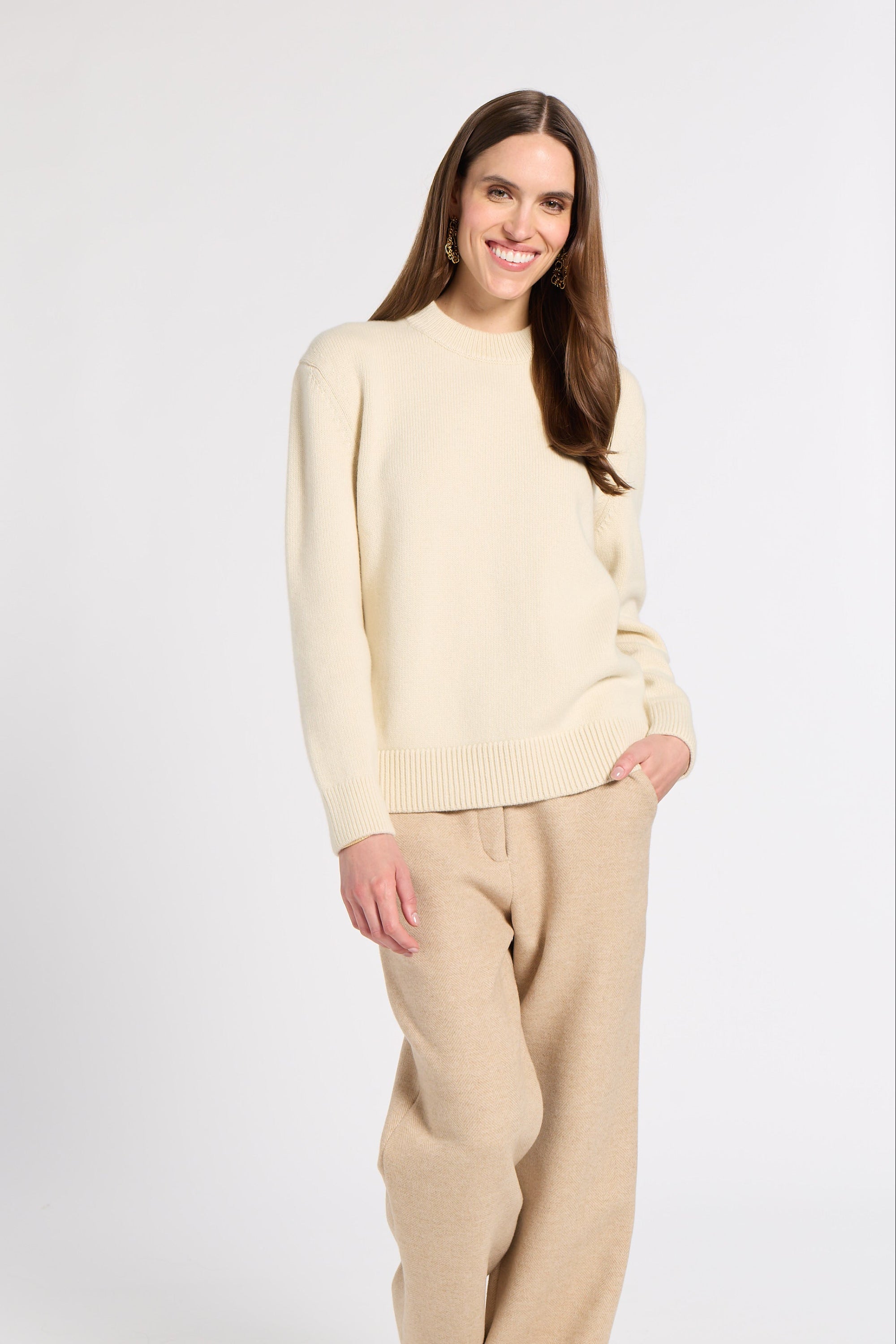 A woman modeling a cream knit crewneck sweater and beige wide-leg trousers against a plain light gray studio background.