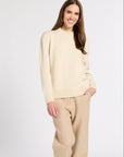 A woman modeling a cream knit crewneck sweater and beige wide-leg trousers against a plain light gray studio background.