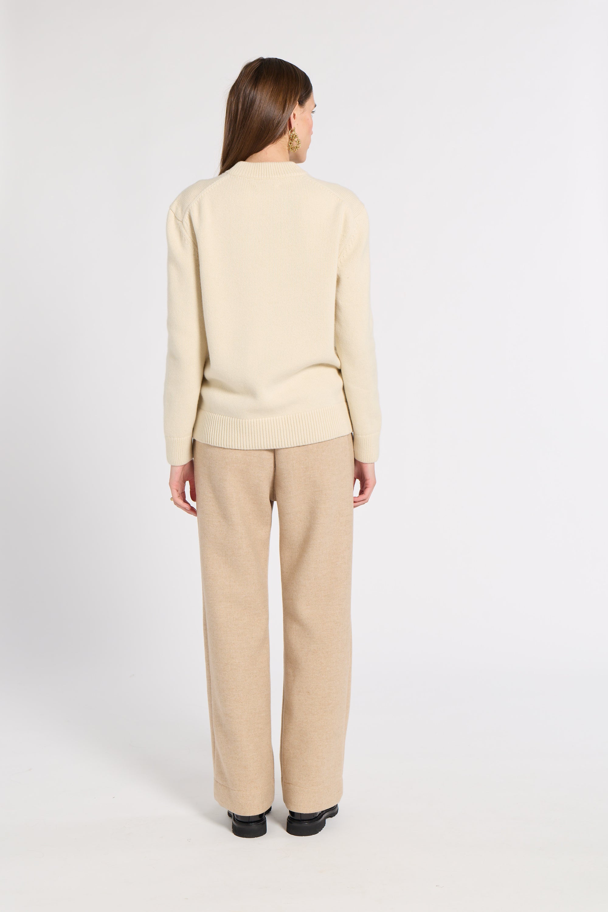 A woman viewed from the back wearing a cream ribbed knit sweater and beige wide-leg trousers against a plain light gray studio background.
