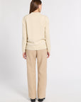 A woman viewed from the back wearing a cream ribbed knit sweater and beige wide-leg trousers against a plain light gray studio background.