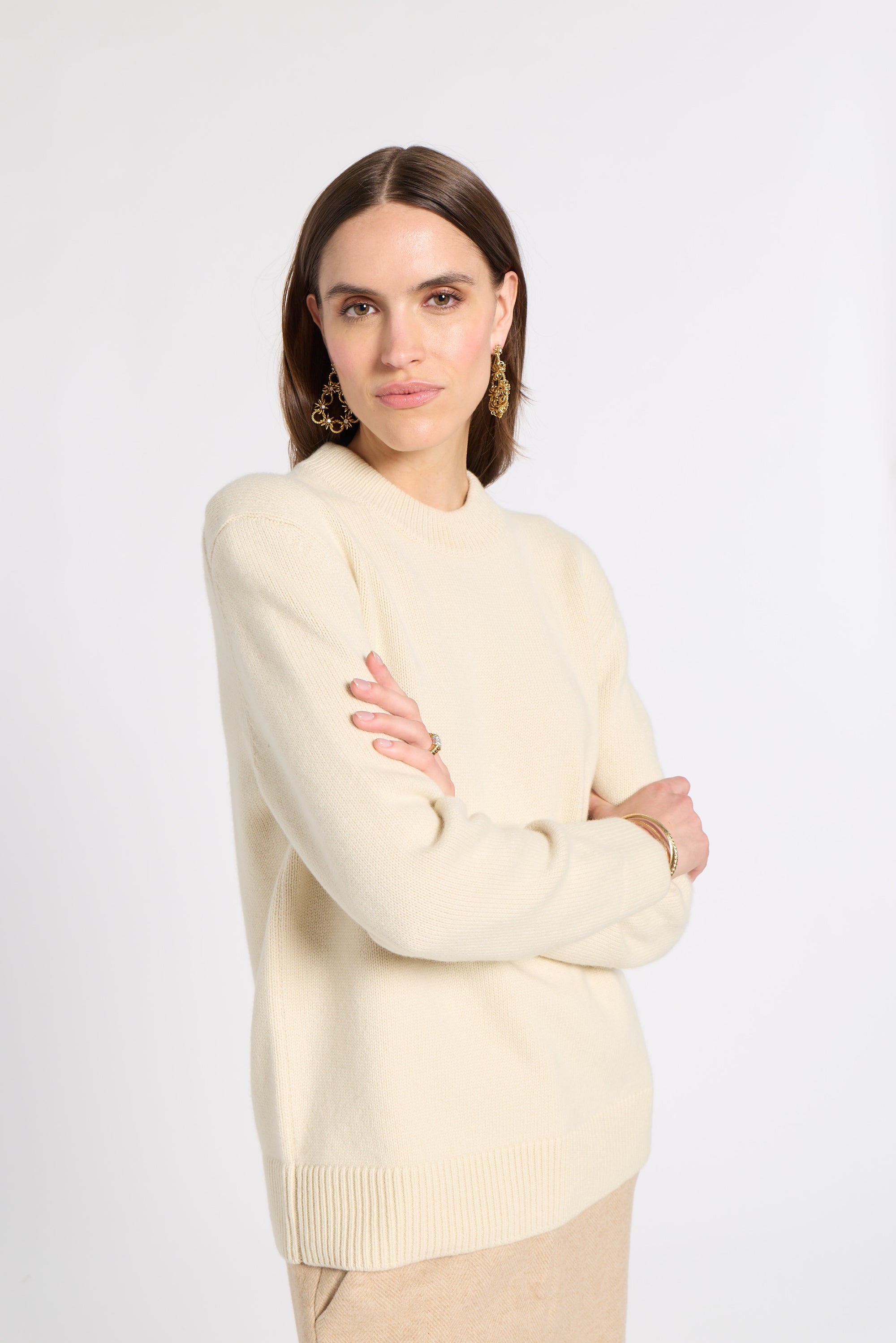 A woman modeling a cream-colored knit sweater with a crew neck and ribbed hem, paired with beige trousers, against a plain light gray studio background.