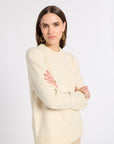 A woman modeling a cream-colored knit sweater with a crew neck and ribbed hem, paired with beige trousers, against a plain light gray studio background.
