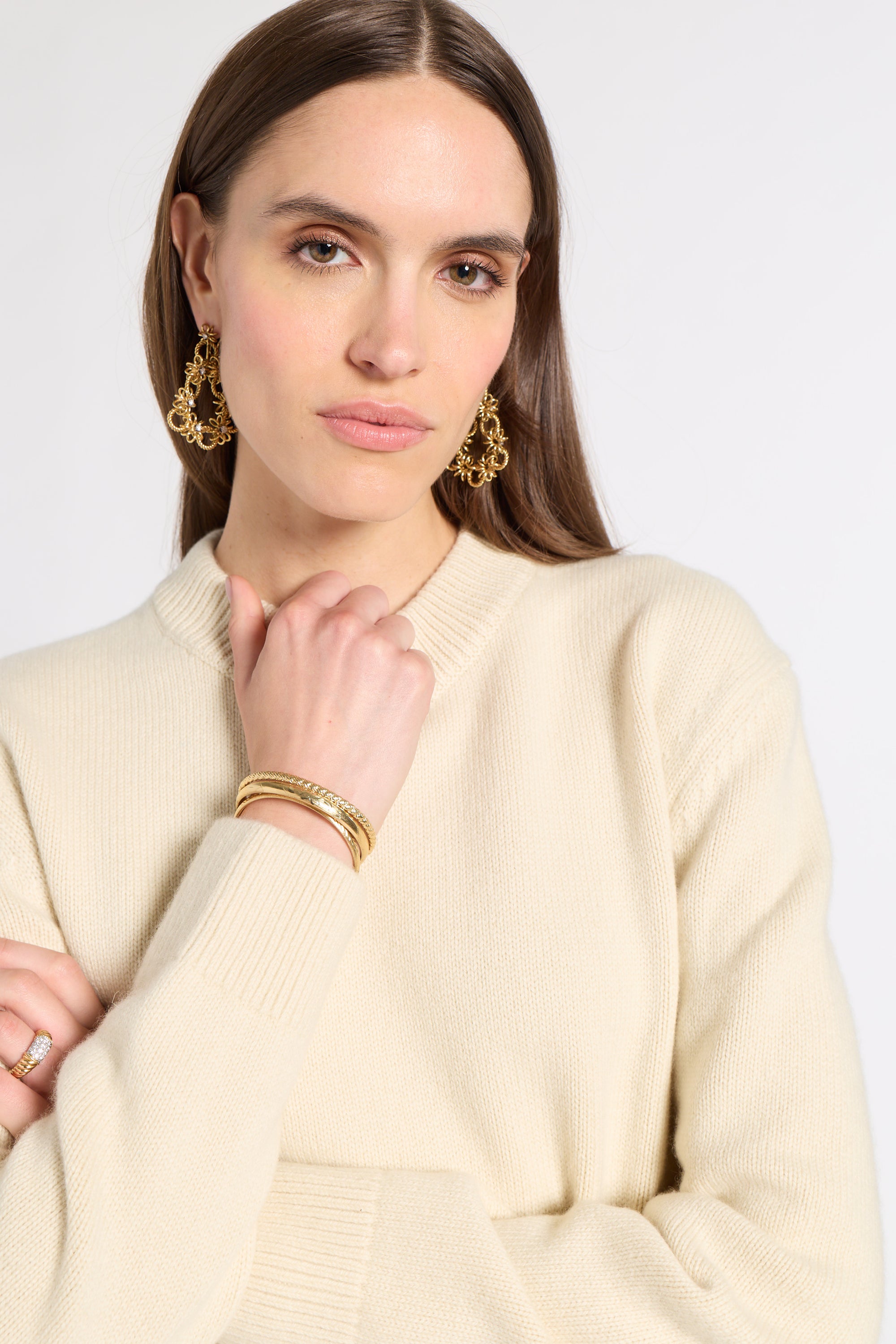 A woman wearing a cream-colored ribbed knit sweater, gold hoop earrings, and stacked gold bracelets, shown against a plain light gray studio background.