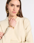A woman wearing a cream-colored ribbed knit sweater, gold hoop earrings, and stacked gold bracelets, shown against a plain light gray studio background.