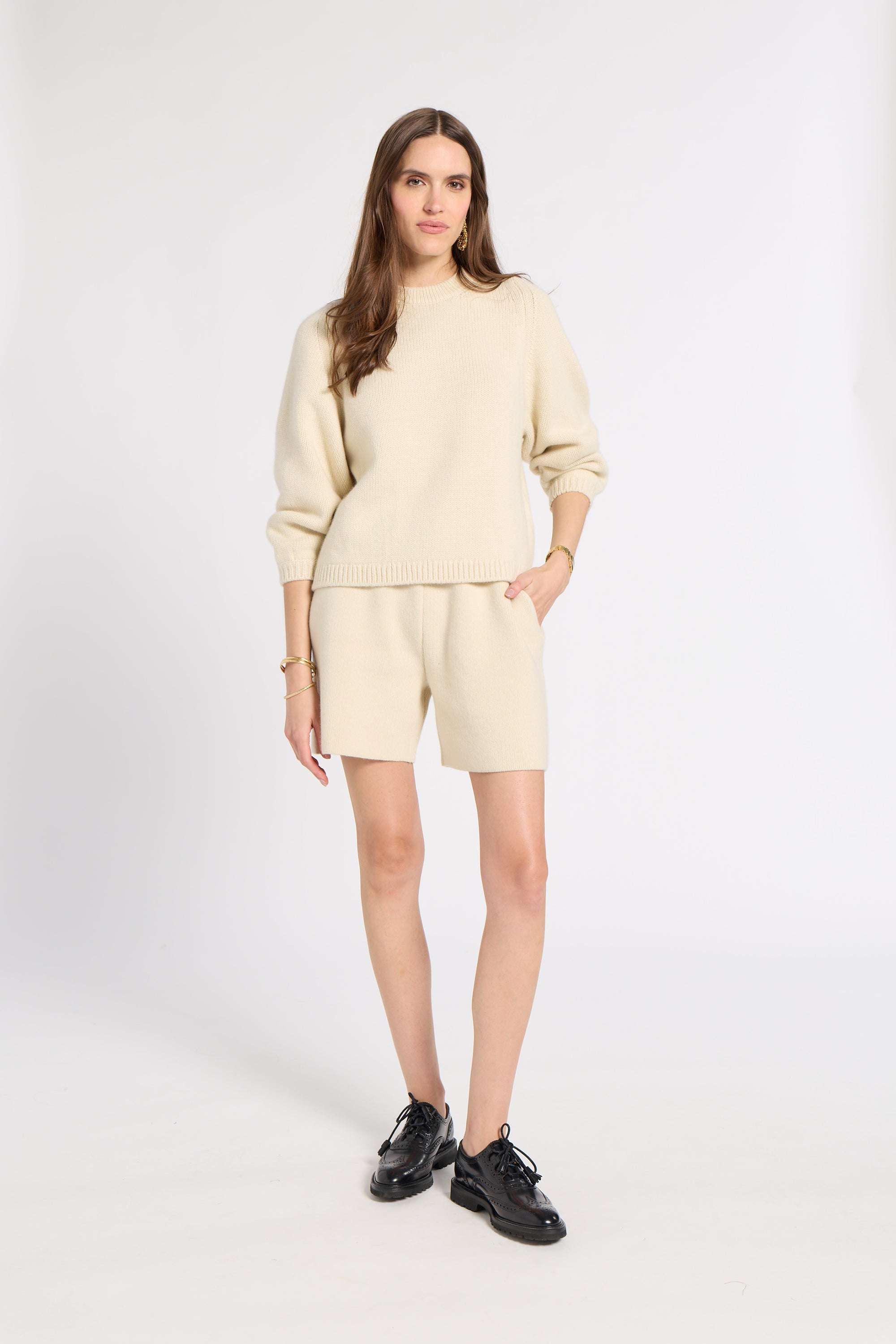 A woman modeling a cream-colored knit sweater and matching shorts set, paired with black leather shoes, against a plain light gray studio background.