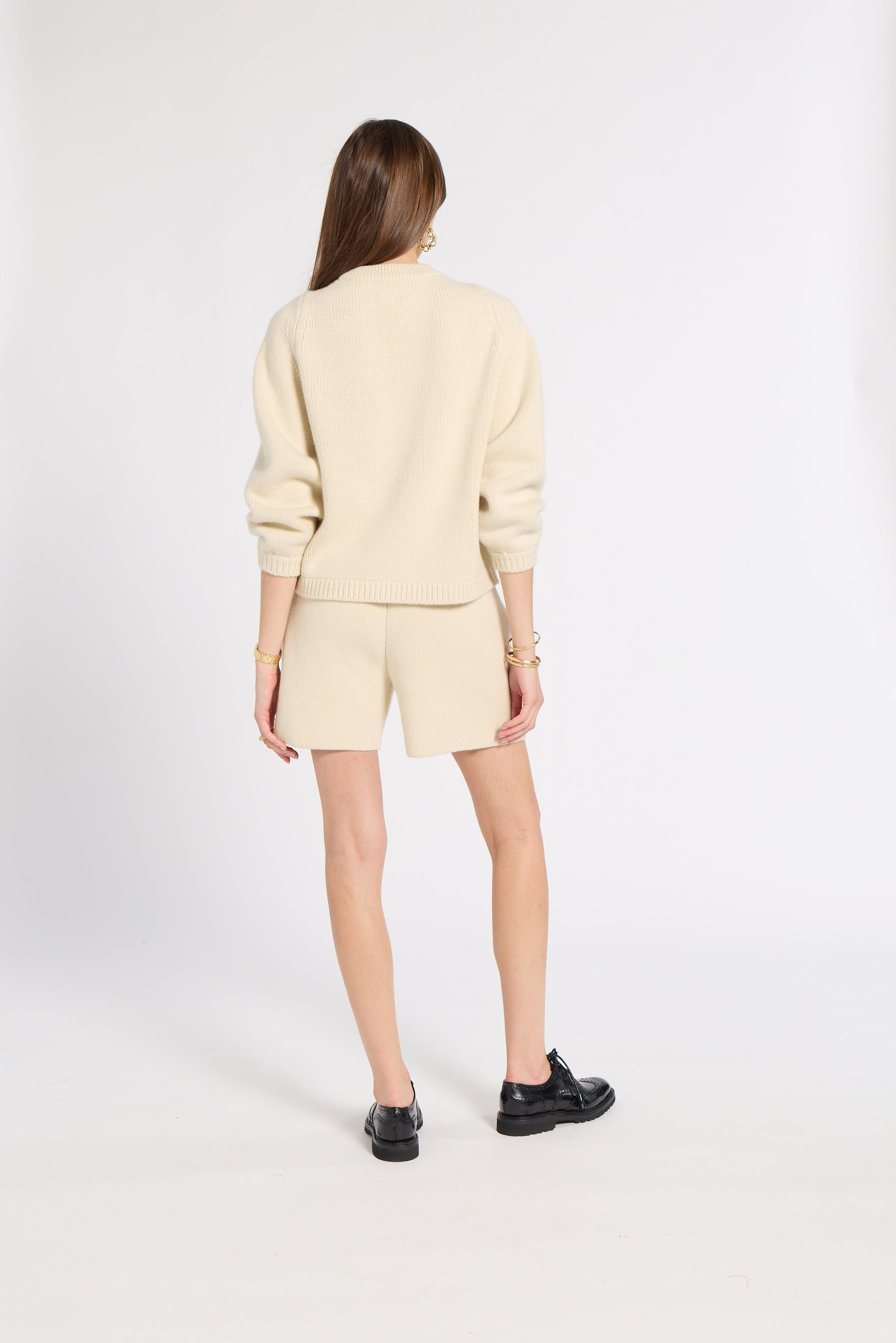 A model is shown from the back wearing a cream-colored knit mock-neck sweater and matching shorts, paired with black leather shoes, against a plain white studio background.