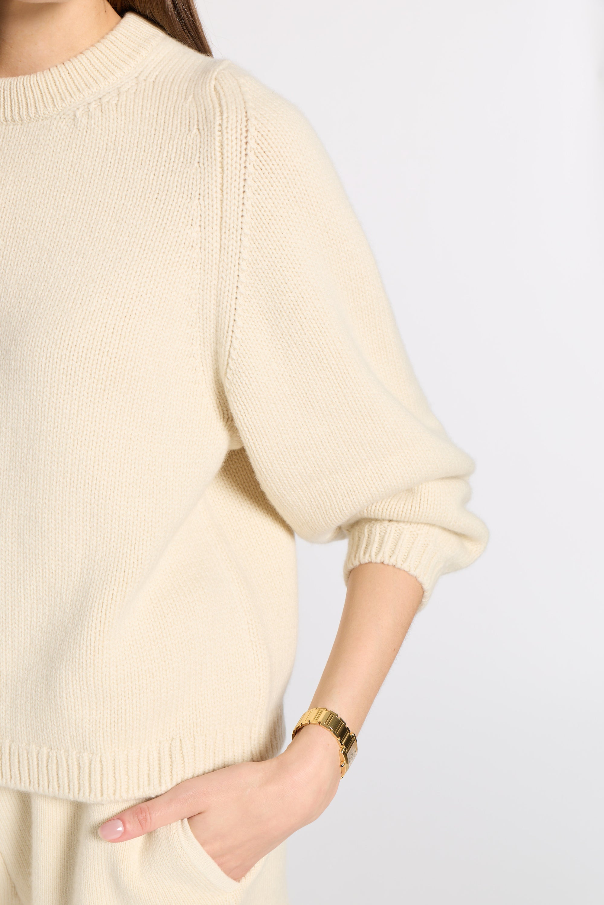 A close-up of a person wearing a cream-colored ribbed knit sweater with a gold watch on their wrist, set against a plain light gray background.