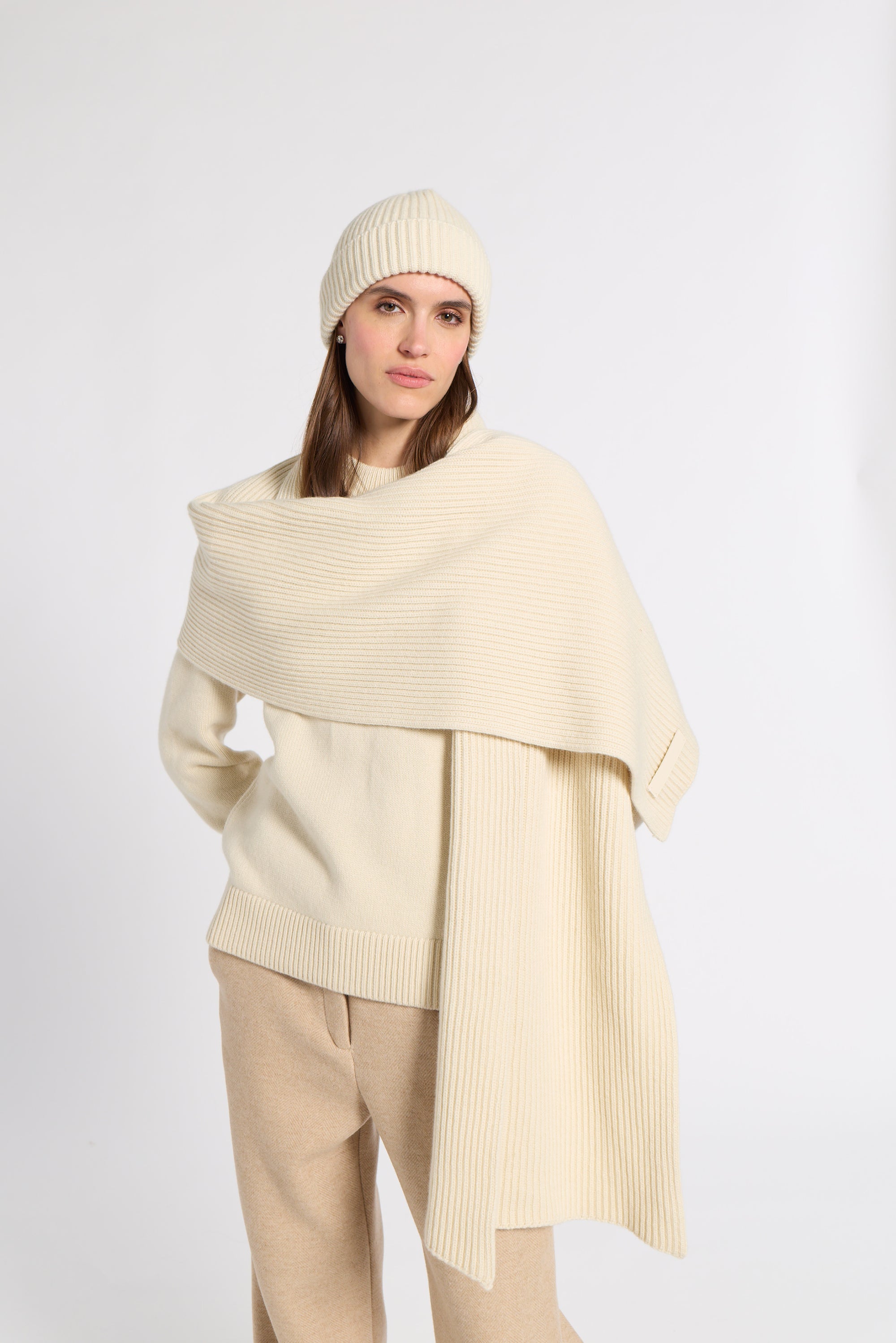 A woman modeling a cream-colored ribbed knit set, featuring a draped poncho-style sweater, a matching ribbed beanie, and beige trousers, shot against a plain light gray studio background.