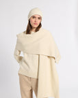 A woman modeling a cream-colored ribbed knit set, featuring a draped poncho-style sweater, a matching ribbed beanie, and beige trousers, shot against a plain light gray studio background.