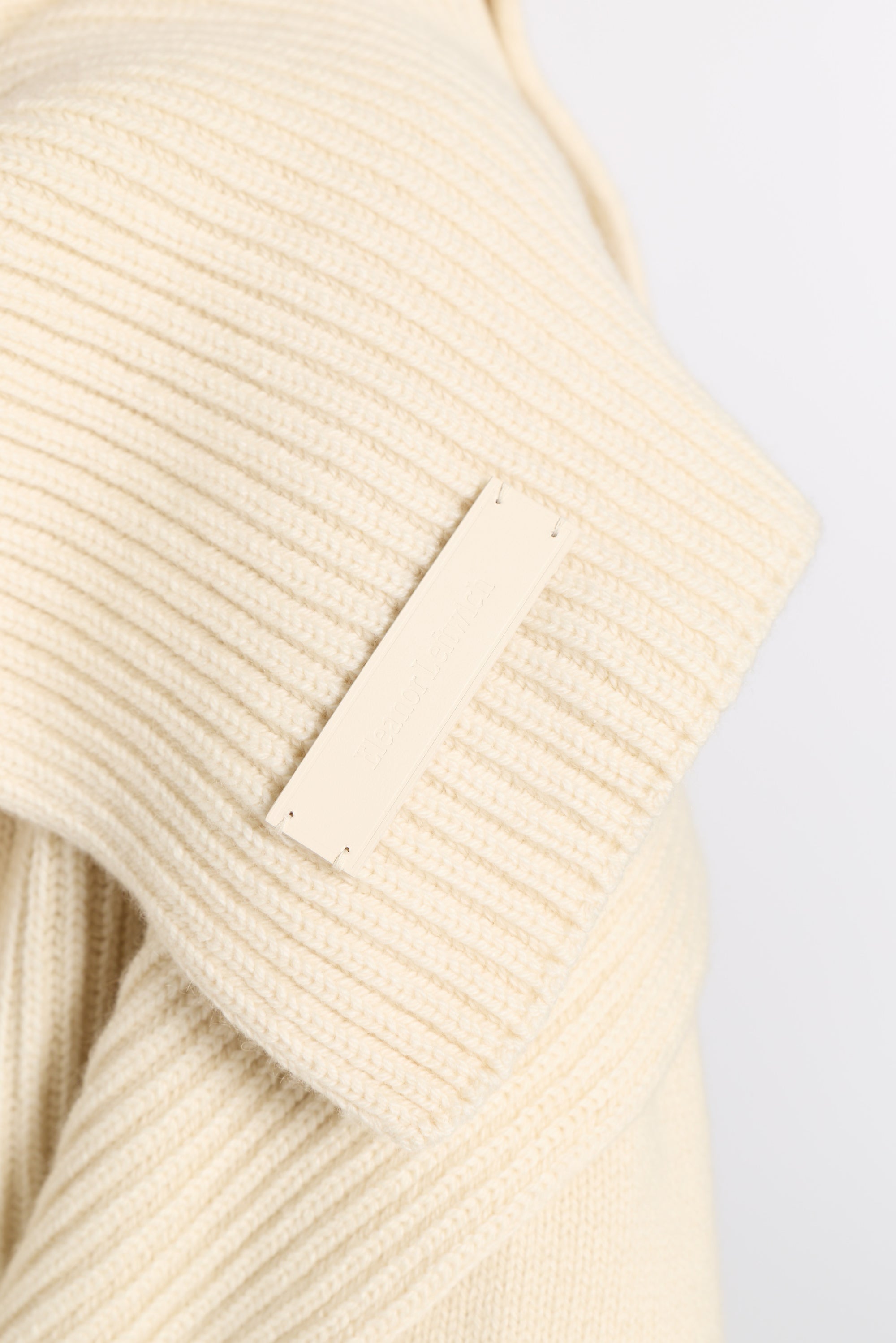 Close-up of a cream-colored ribbed knit garment with a blank rectangular fabric label sewn onto the shoulder.