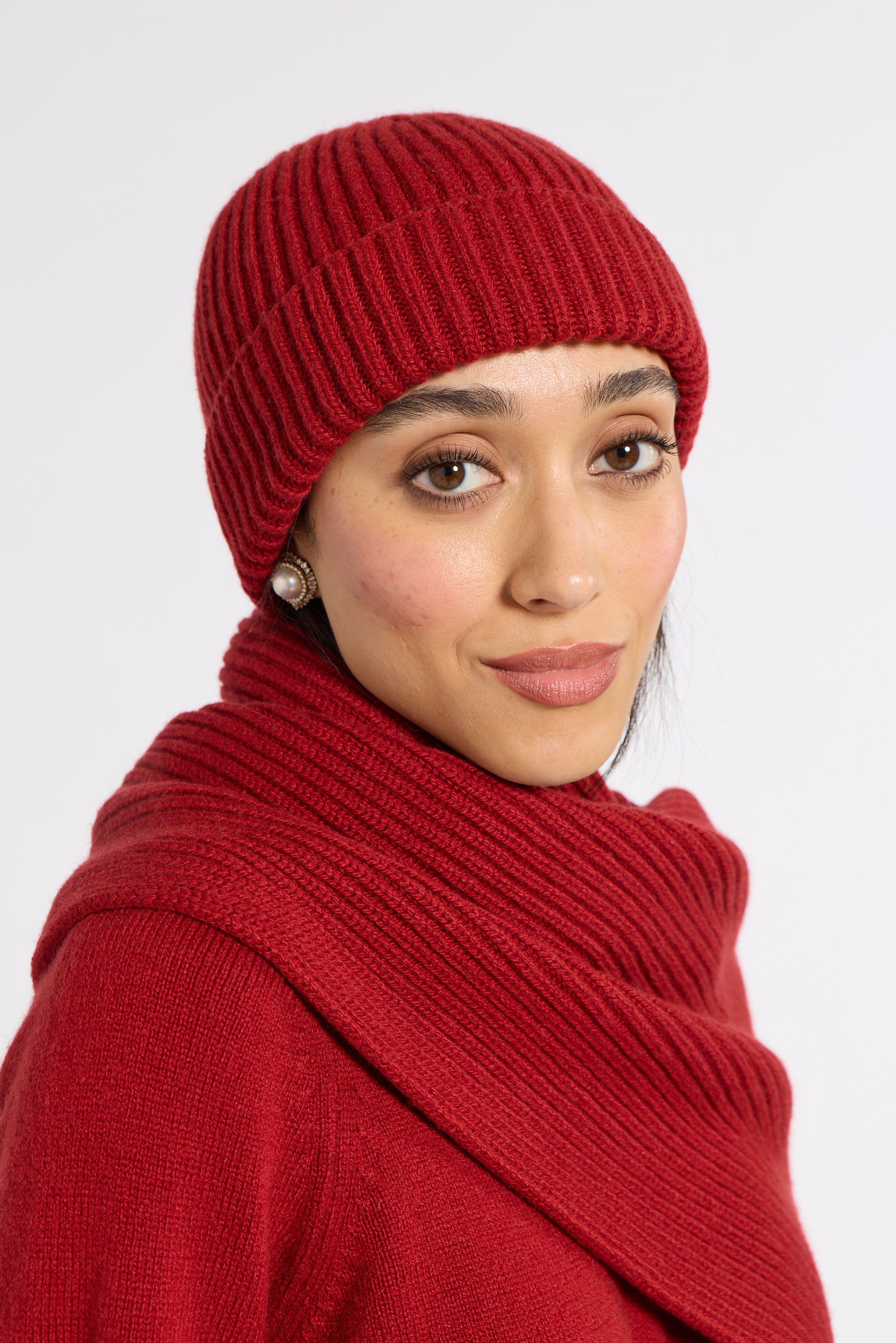 A woman wearing a matching red ribbed knit beanie and thick cowl neck scarf, photographed against a clean light gray studio background.