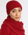 A woman wearing a matching red ribbed knit beanie and thick cowl neck scarf, photographed against a clean light gray studio background.