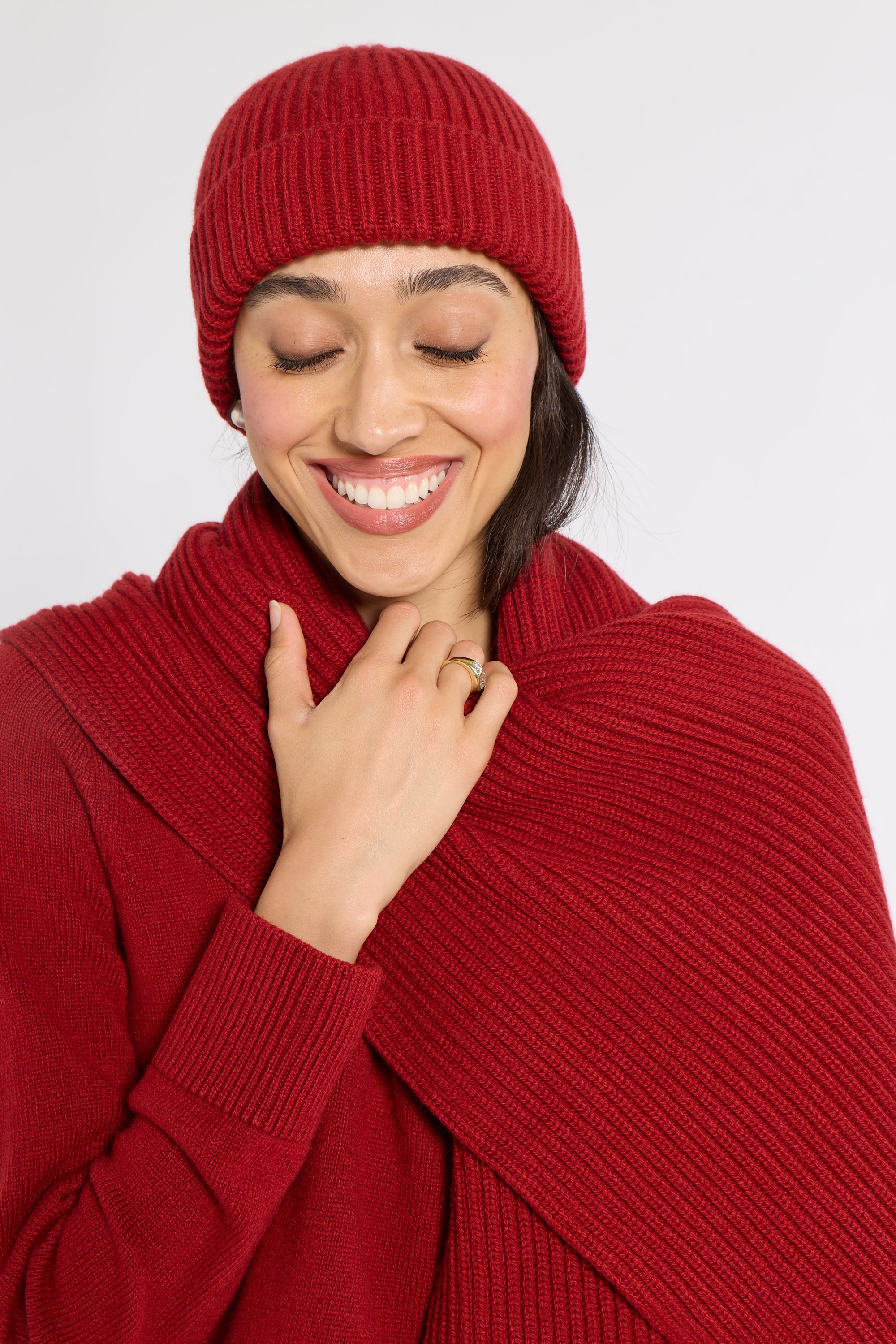 A smiling woman wearing a matching ribbed knit beanie and a thick ribbed knit wrap or scarf in a deep red color, posed against a light gray studio background.